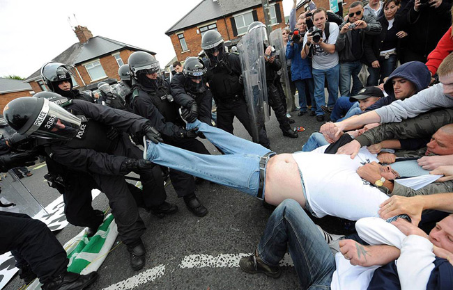Police and rioters come together to help fat man out of trousers.