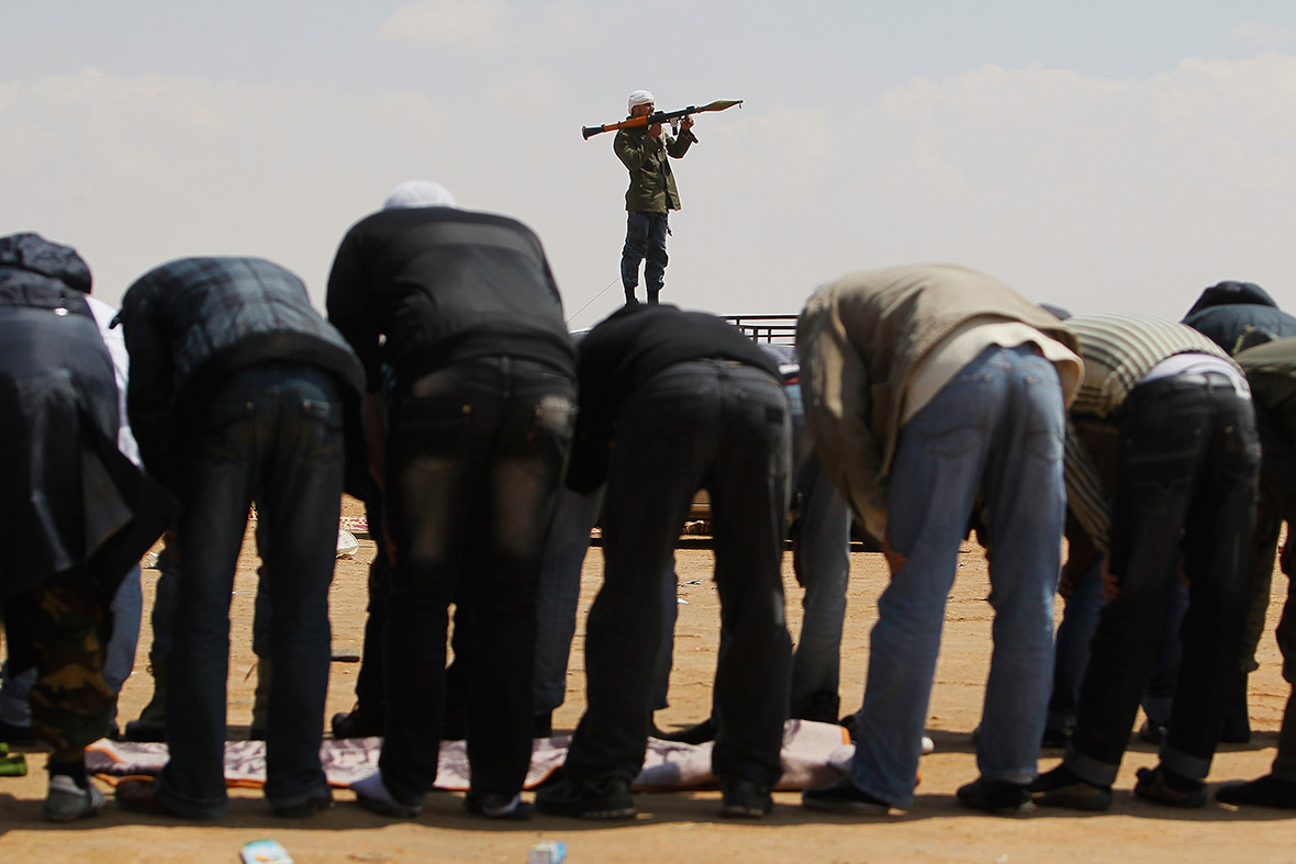 April 8, 2011: Rebels pray while a comrade stands with a rocket propelled grenade launcher on the western edge of Ajdabiyah, Libya