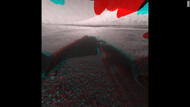 This image is a 3-D view in front of NASA's Curiosity rover. The anaglyph was made from a stereo pair of Hazard-Avoidance Cameras on the front of the rover. Mount Sharp, a peak that is about 3.4 miles high, is visible rising above the terrain, though in one "eye" a box on the rover holding the drill bits obscures the view. 