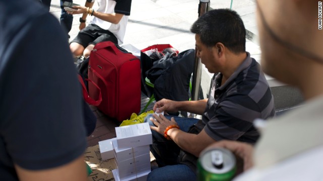 Resellers sit on cheap plastic chairs with their open suitcases beside them, stacked up with white boxes. 