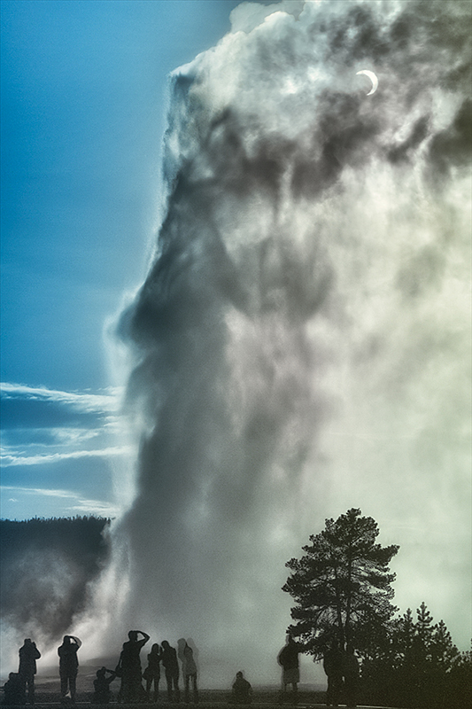 Eclipse and Old Faithful by Robert Howell (USA). Visitors witness the Old Faithful geyser in Yellowstone National Park erupt as the Moon partially eclipses the Sun. The scene captures a sense of awe set against blue sky and white geyser steam, as the onlookers strain to see the joining of these two phenomena – one geological and one astronomical.