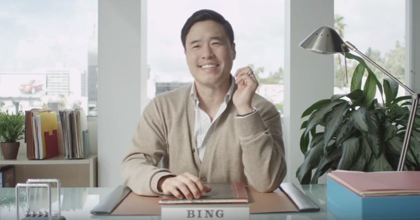 A smiling Asian man in his 30s sits behind a desk in an airy, light office with a large window behind him. A nameplate on his desk reads 'BING'.