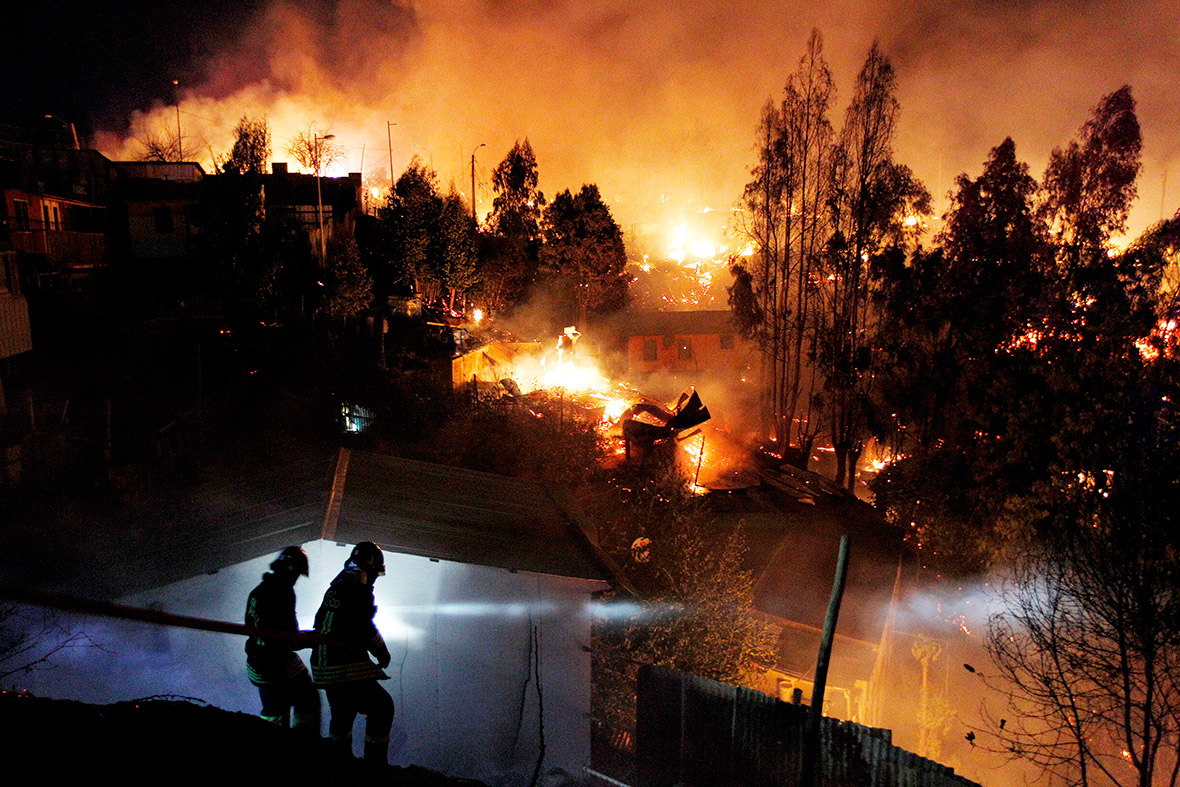 Two firefighters investigate an area while a huge fire consumes hillside properties