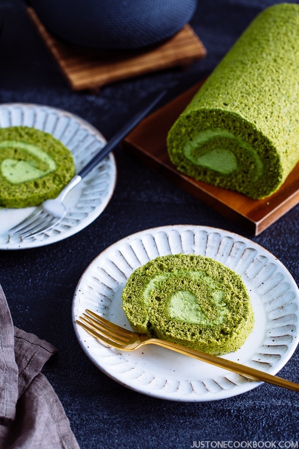 A slice of matcha swiss roll on the white plates with uncut roll cake on the back.
