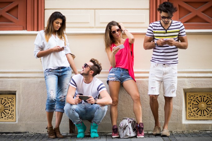 Group of four people in the city standing (and one is crouching) next to a building and operatng their mobile phones. Looks like they are testing mobile applications or social media.