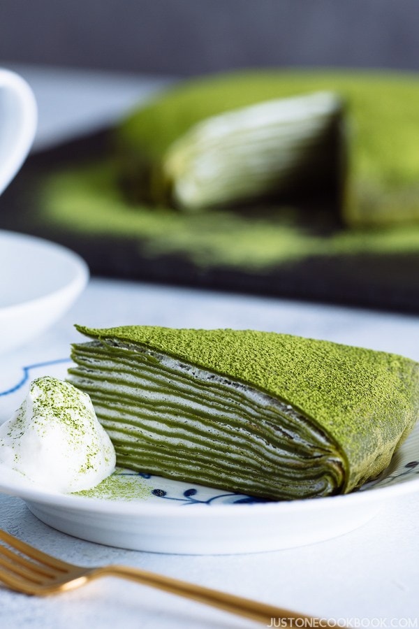 A slice of matcha mille crepe on a Royal Copenhagen plate with fresh whipped cream on top.