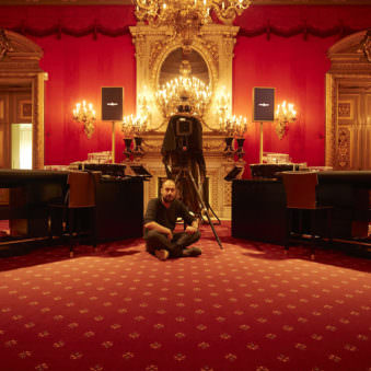 Portrait eines sitzenden jungen Mannes in einem roten, barocken Zimmer mit Großformatkamera im Bild.