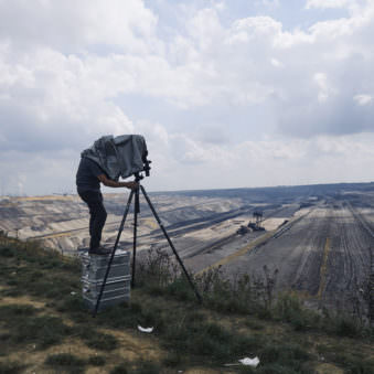 Person steht auf einer Kiste in einer Landschaft mit viel Himmel und schaut durch eine Großfomratkamera auf einem Stativ.