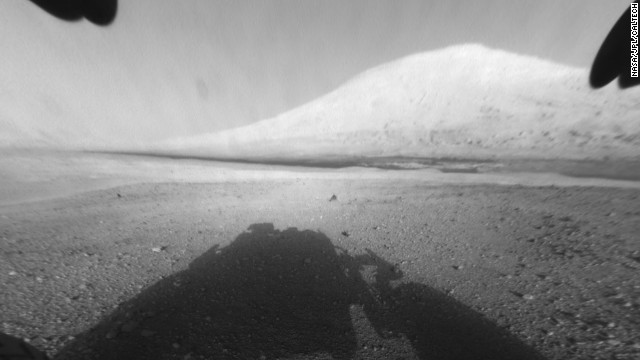 This image shows Curiosity's main science target, Mount Sharp. The rover's shadow can be seen in the foreground. The dark bands in the distances are dunes. 