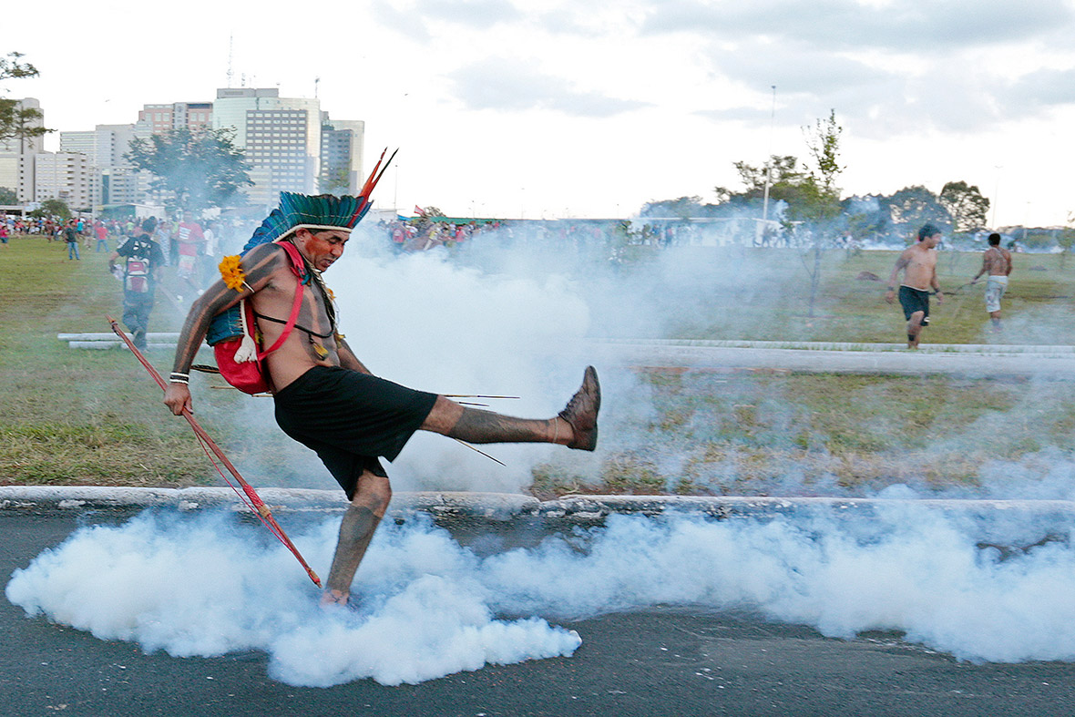 An indigenous Brazilian kicks a tear gas grenade back at riot police blocking protesters from approaching the Mane Garrincha soccer stadium in Brasilia